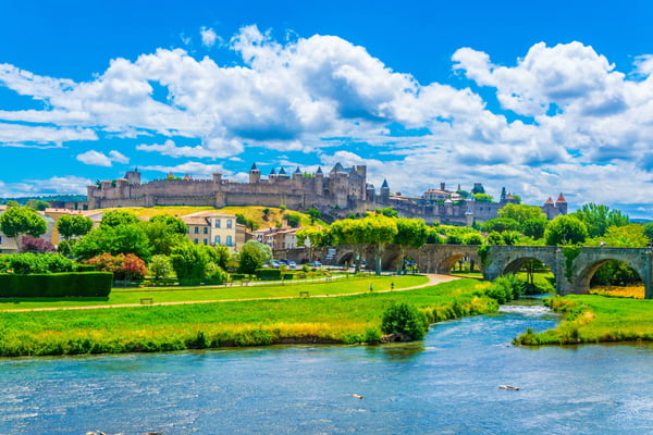 Blick auf die mittelalterliche Festungsstadt Carcassonne mit Stadtmauern und Türmen, davor Fluss, Grünflächen und eine Steinbrücke.
