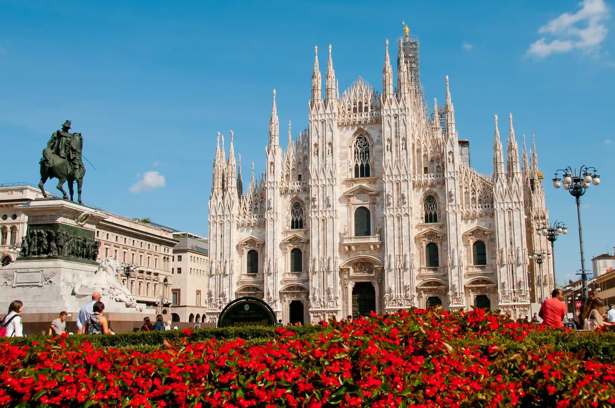 Façade gothique du Dôme de Milan avec de nombreuses flèches sous un ciel bleu et parterre de fleurs au premier plan
