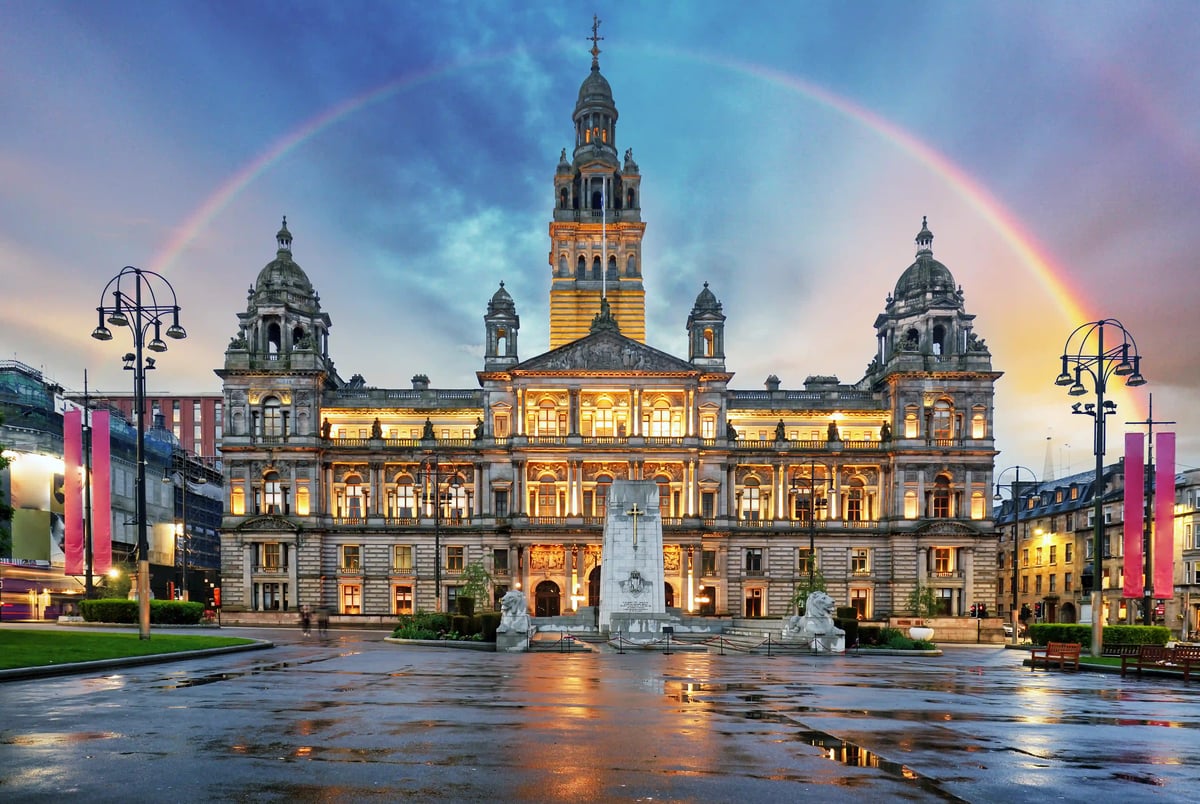 Beleuchtetes historisches Rathaus bei Dämmerung mit Regenbogen über dem Gebäude und spiegelnder nasser Platzfläche