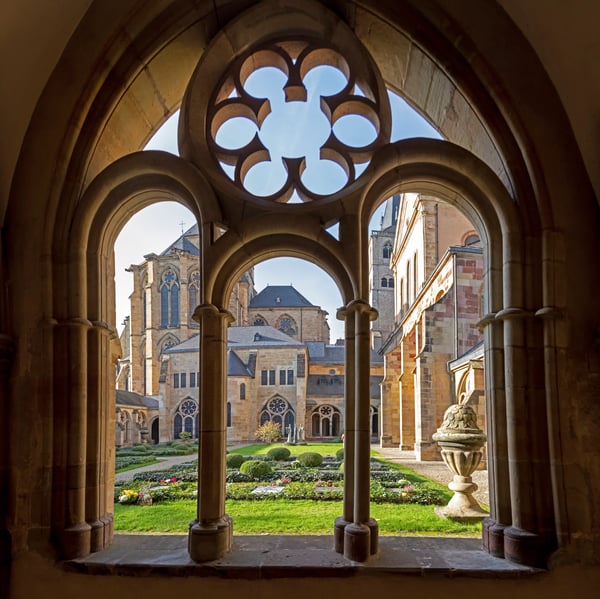 Blick durch steinerne Rundbögen in einen Klostergarten mit Dom und historischen Gebäuden in Trier