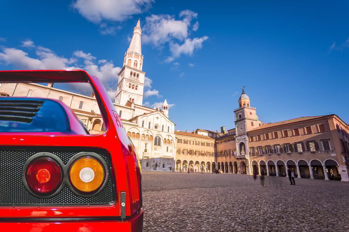 Historischer Domplatz in Modena mit romanischem Dom und Glockenturm unter blauem Himmel, im Vordergrund das Heck eines roten Sportwagens