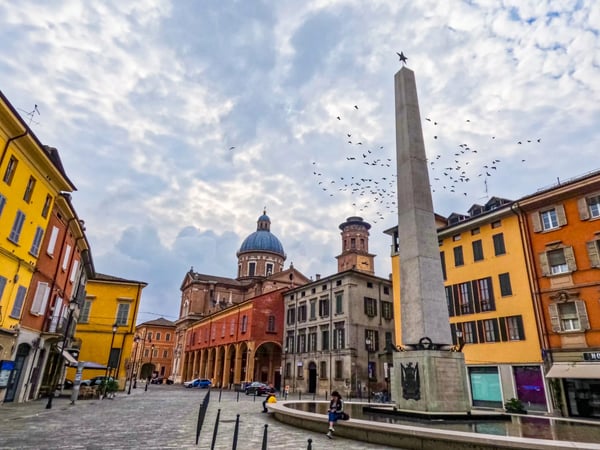 Weiter Platz mit historischem Obelisk, farbigen Häuserfassaden und barocker Kirche unter bewölktem Himmel