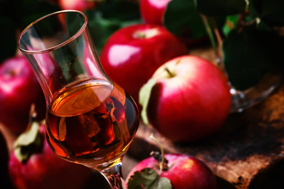 Glas mit bernsteinfarbenem Calvados, umgeben von roten Äpfeln auf rustikalem Holztisch