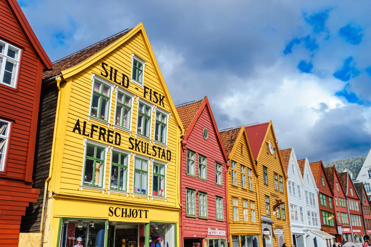 Reihe farbiger Holzhäuser in Bergen, im Vordergrund ein gelbes Haus mit Schildern, dramatischer Himmel mit Wolken.