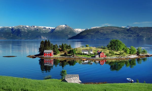 Ruhiger Fjord mit kleiner Insel und roten Häusern, Spiegelungen im Wasser, dahinter Berge mit Schneefeldern unter blauem Himmel.