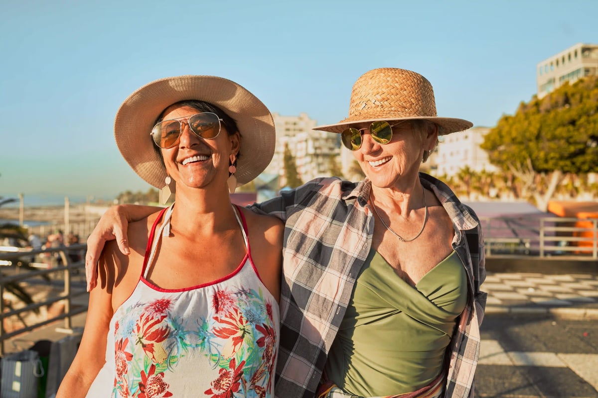 Zwei Frauen mit Hüten und Sonnenbrillen stehen lächelnd an einer Promenade am Meer