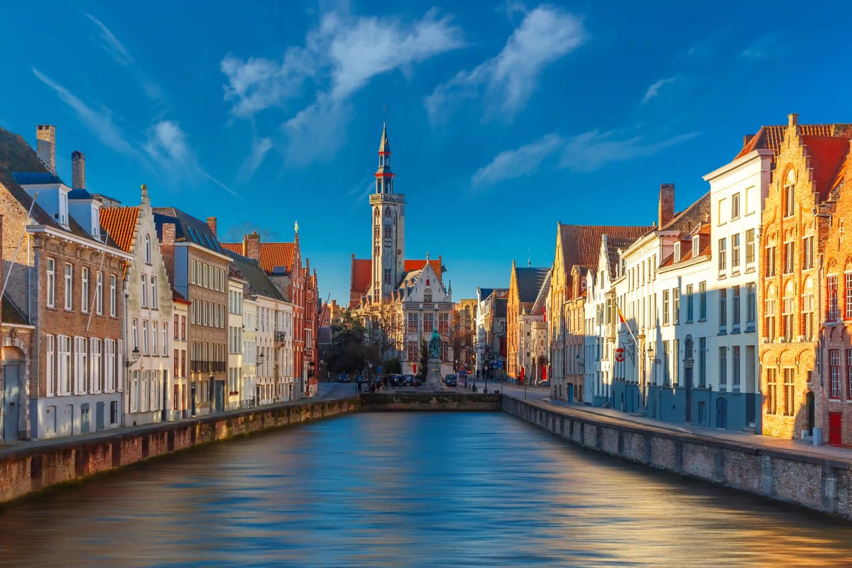 Vue le long dun canal à Bruges, avec rangées de maisons historiques et le beffroi au fond sous un ciel bleu