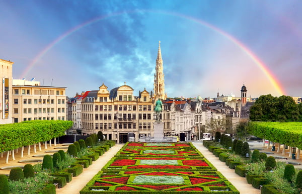 Symmetrische Gartenanlage in Brüssel mit Blumenbeeten, Reiterstatue und Blick auf die Stadtsilhouette, darüber ein Regenbogen