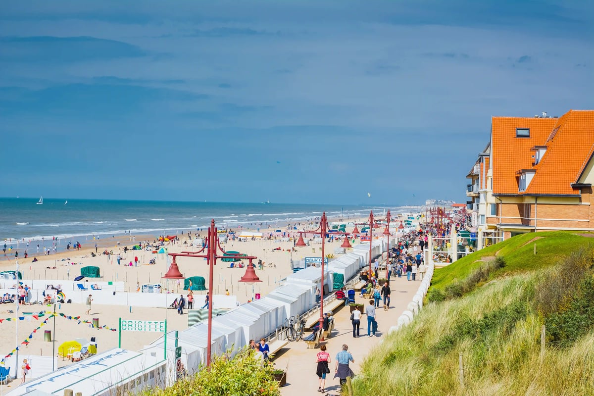 Promenade animée sur la côte belge de la mer du Nord, avec plage de sable, cabines, promeneurs et maisons aux toits rouges sur la digue