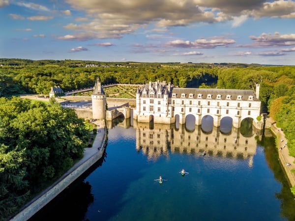 Luftaufnahme von Schloss Chenonceau, das mit mehreren Bögen über einen Fluss gebaut ist, umgeben von Wald und Gartenanlagen.