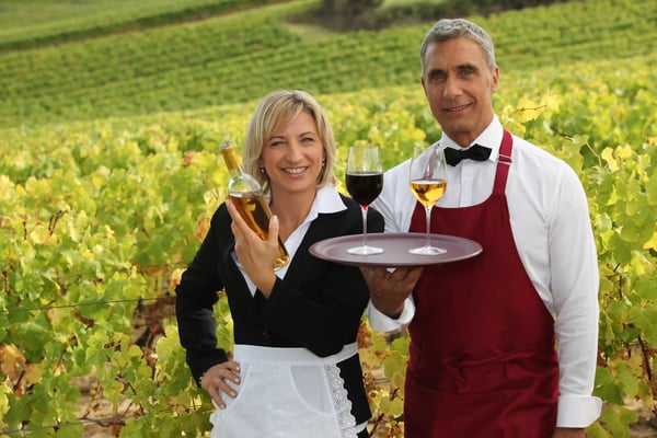 Zwei Servicekräfte im Weinberg halten eine Flasche und ein Tablett mit zwei Weingläsern vor grünen Reben.