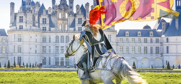 Reiter in historischer Kleidung auf einem weissen Pferd vor einem prachtvollen Schloss mit wehender Fahne im Hintergrund