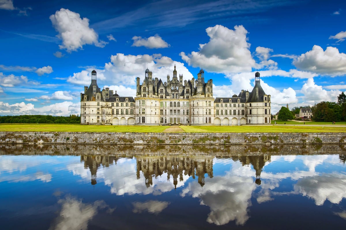 Schloss Chambord mit vielen Türmen steht am Ufer eines ruhigen Flusses, davor zwei Schwäne auf dem Wasser unter blauem Himmel.