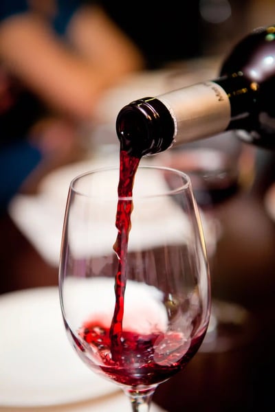 Rotwein fliesst aus einer Flasche in ein Weinglas, der Hintergrund ist unscharf und zeigt eine Person am Tisch.