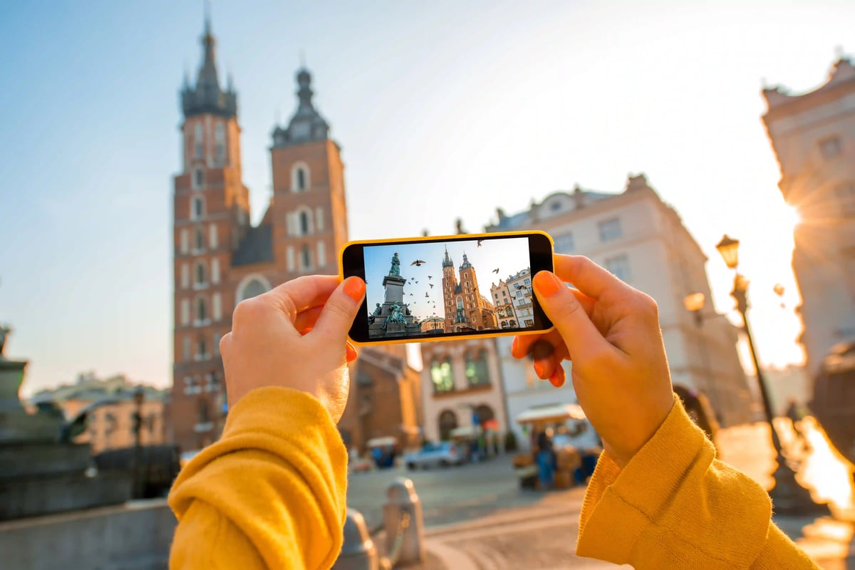 Person fotografiert den Hauptplatz von Krakau mit Smartphone vor historischen Tuerme und Gebäuden bei warmem Licht