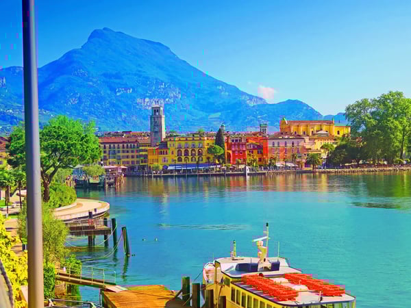 Blick auf die Uferpromenade von Riva del Garda mit bunten Häusern, Booten und Bergen im Hintergrund