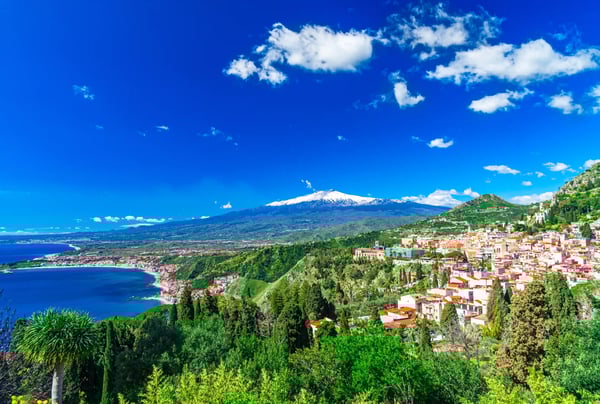 Blick über grüne Hügel und die Küste von Taormina mit dem schneebedeckten Ätna unter blauem Himmel