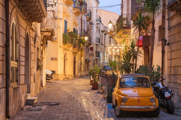 Schmale Altstadtgasse in Siracusa mit Balkonen warmem Licht und parkendem Kleinwagen