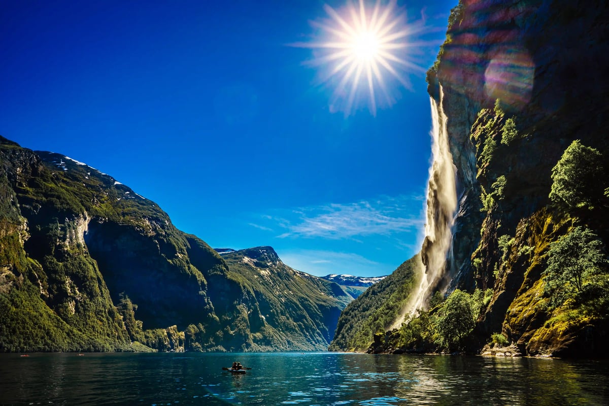 Geirangerfjord mit steilen grünen Bergen, Wasserfall an der Felswand und glitzerndem Wasser unter strahlender Sonne