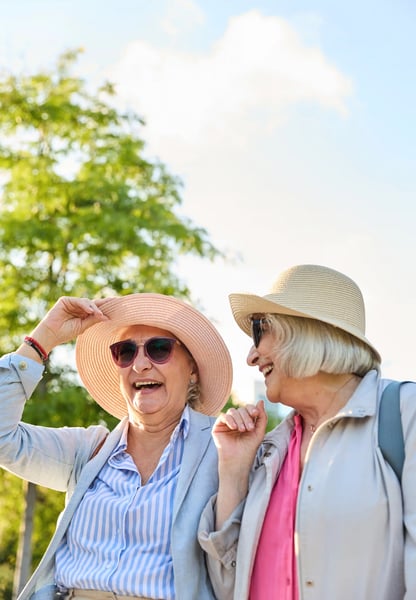 Zwei ältere Frauen mit Sonnenhüten lachen und unterhalten sich bei einem Spaziergang im Grünen
