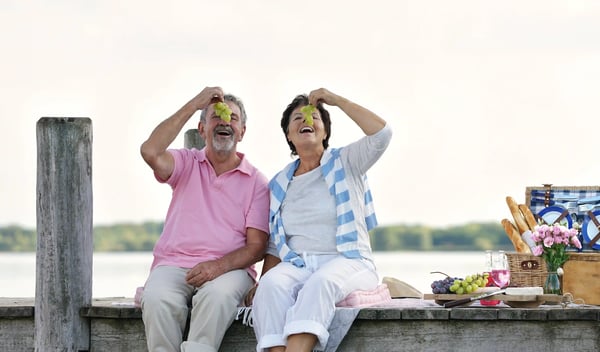 Älteres Paar sitzt am Steg und hält lachend Trauben in den Händen bei einem Picknick
