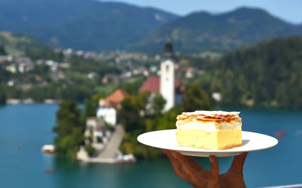 Bleder Cremeschnitte auf einem Teller im Vordergrund, im Hintergrund der Bleder See mit Insel und Kirche