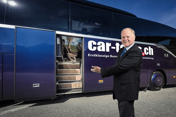 Reiseleiter steht lächelnd vor einem modernen Reisebus und zeigt einladend auf den geöffneten Einstieg mit bequemen Sitzen im Inneren