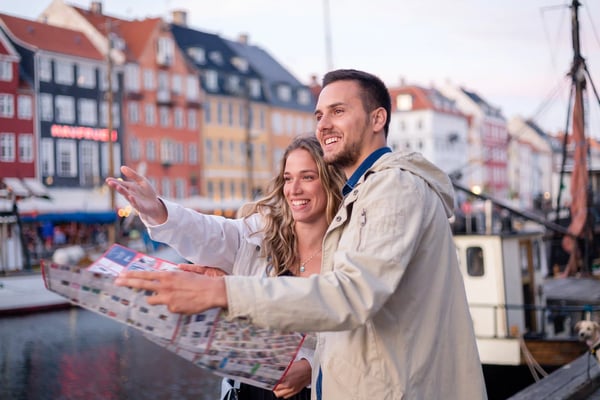 Junges Paar am Hafen von Kopenhagen betrachtet einen Stadtplan und zeigt lächelnd in Richtung der bunten Häuser im Hintergrund