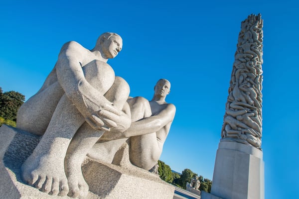 Granitskulpturen sitzender Figuren und hoher Monolith im Vigeland-Park in Oslo vor blauem Himmel