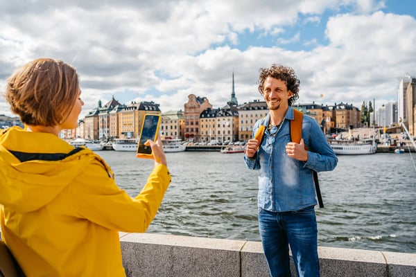 Frau fotografiert einen lächelnden Mann mit Rucksack an der Uferpromenade von Stockholm vor historischer Stadtkulisse