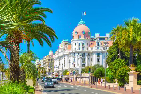 Strassenszene in Nizza mit Palmen, historischem Hotelgebäude, Autos und Passanten unter blauem Himmel