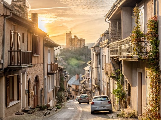 Schmale Dorfstrasse in Najac bei Abendlicht, Steinhäuser und parkierte Autos, Festung auf dem Hügel im Hintergrund