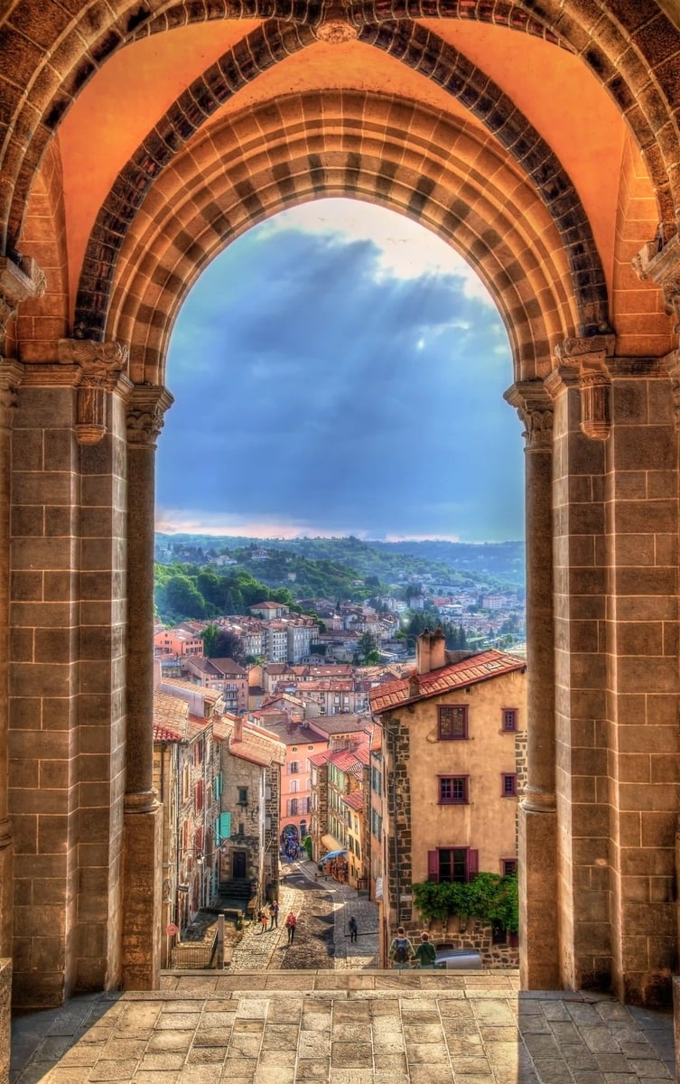 Blick durch einen grossen Steinbogen auf die Altstadt mit roten Dächern, Sonnenstrahlen brechen durch dunkle Wolken