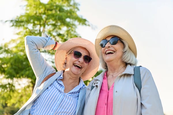 Zwei ältere Frauen mit Sonnenhüten und Sonnenbrillen lachen im Freien vor grünem Hintergrund