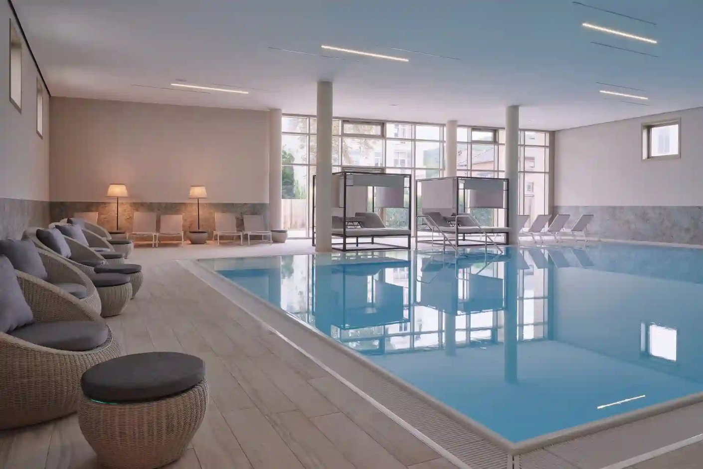 Helles Hallenbad mit rechteckigem Pool, grossen Fenstern, Liegen und Sitzmöbeln in ruhiger Spa-Atmosphäre.