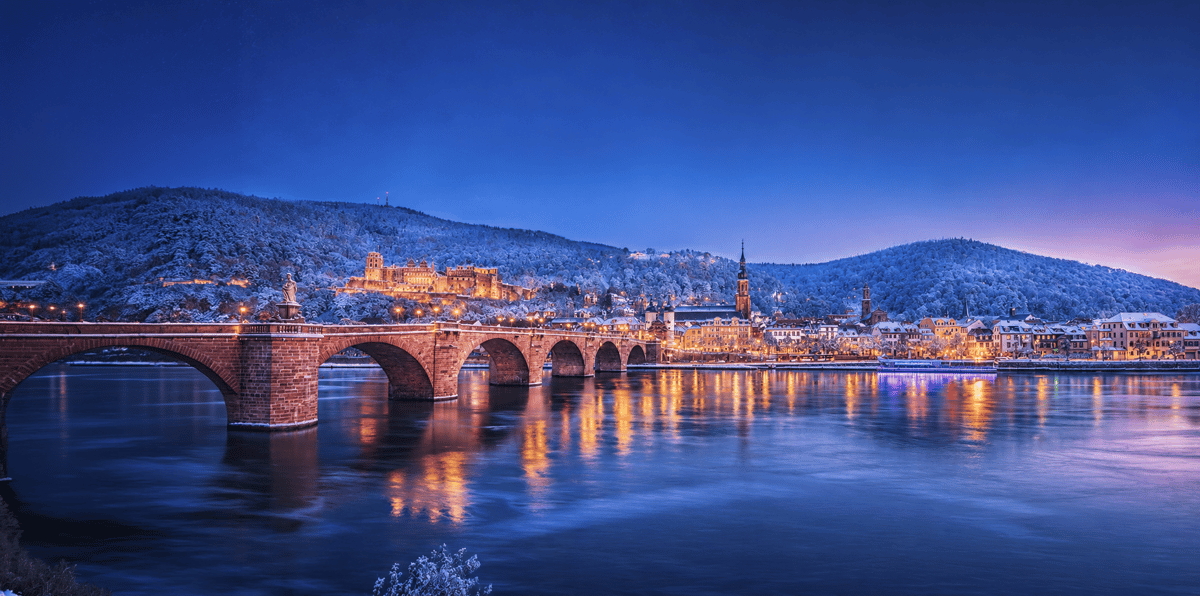 Beleuchtetes Heidelberger Schloss über der Altstadt und dem Neckar in der Abenddämmerung.