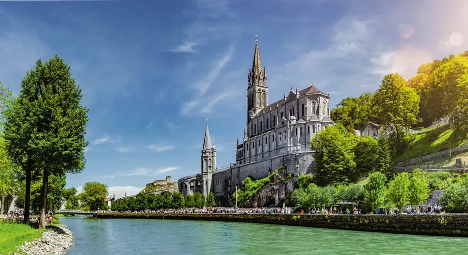 Wallfahrtsbasilika von Lourdes am Fluss Gave mit Pilgerweg und grüner Parklandschaft.