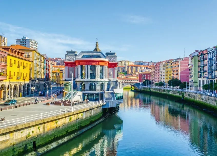 Markthalle am Fluss Nervión in Bilbao, umgeben von historischen Gebäuden und Uferpromenade.