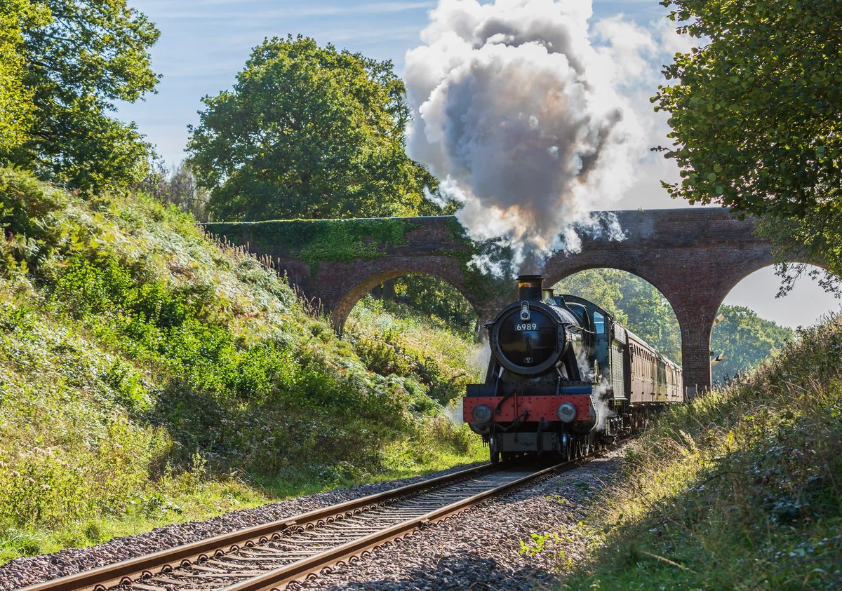 Schwarze Dampflokomotive fährt unter einer gemauerten Bogenbrücke durch grüne Landschaft und stösst dichten weissen Dampf aus