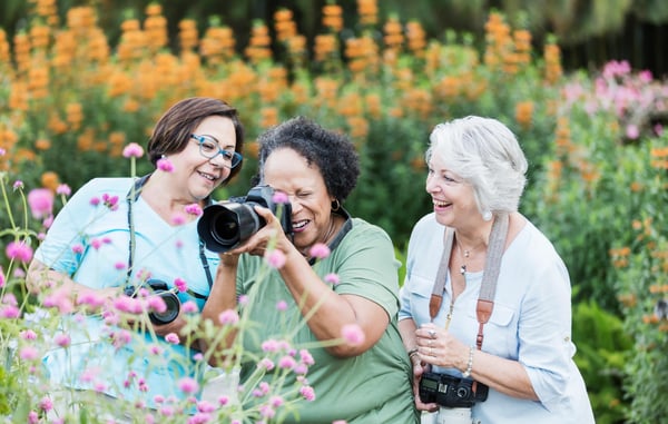Drei ältere Frauen fotografieren lachend Blumen in einem Garten, umgeben von bunten Blüten