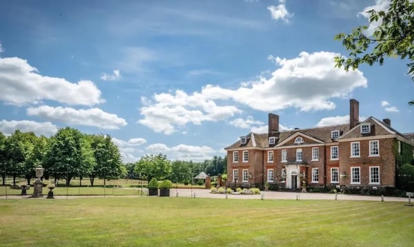 Grosses rotes Backsteingutshaus mit weissem Eingang, umgeben von gepflegtem Rasen und altem Baumbestand unter blauem Himmel