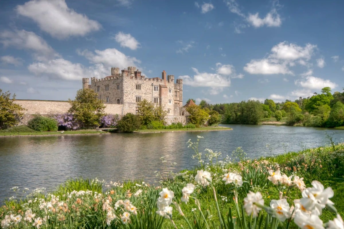 Historisches Steinschloss mit Zinnen am Ufer eines ruhigen Sees, umgeben von Grünanlagen und blühendem Garten