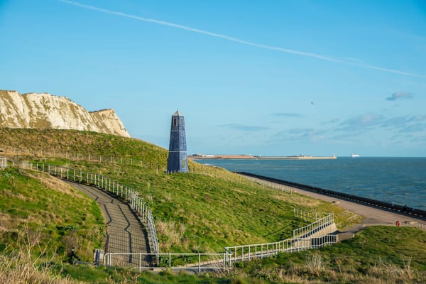 Grüne Küstenlandschaft mit Spazierweg, kleinem Leuchtturm und Blick auf das Meer unter klarem Himmel