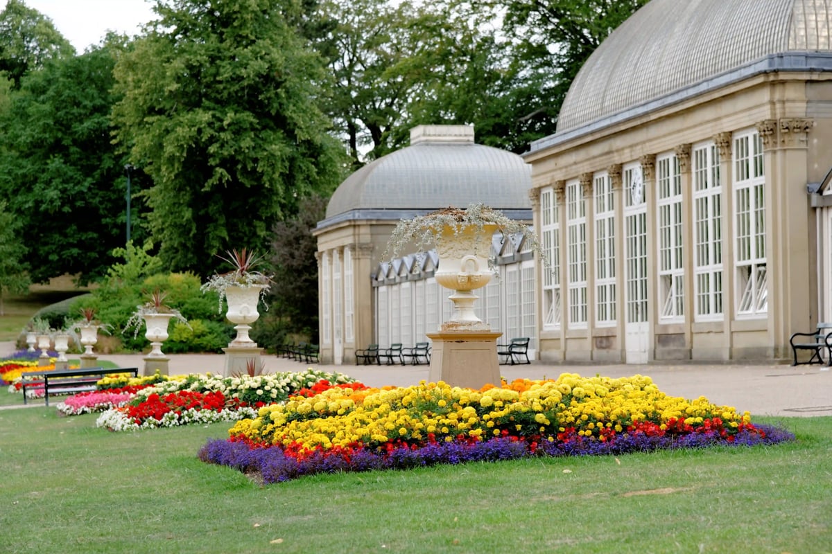 Blumenbeete mit bunten Blueten vor historischer Gewächshausarchitektur in einer gepflegten Parkanlage
