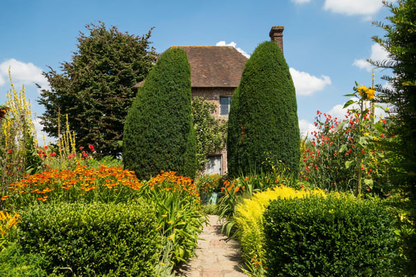 Schmaler Gartenweg führt zwischen dicht geschnittenen Hecken und farbenfrohen Blumen zu einem historischen Haus