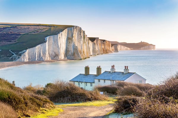 Weisse Kreidefelsen ragen steil aus dem Meer, im Vordergrund ein kleines Haus und ein Küstenpfad