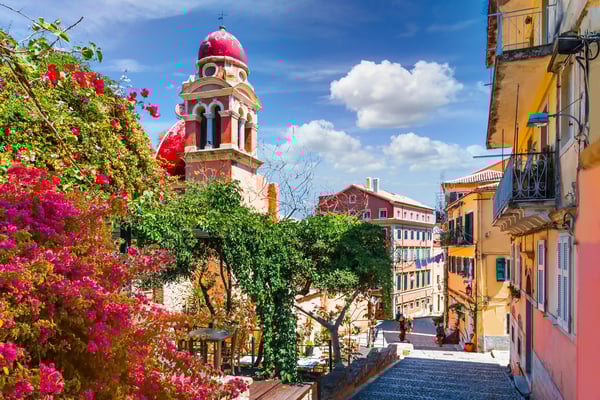 Gasse in der Altstadt von Korfu mit bunten Häusern, roter Kirchturmkuppel, Blumen und Treppenweg
