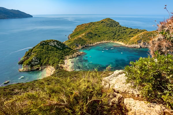 Türkisfarbene Badebucht auf Korfu mit grün bewachsenen Hügeln, Sandstrand und Booten im Wasser
