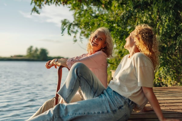 Zwei Frauen sitzen entspannt auf einem Holzsteg am See, lachen miteinander und geniessen die Natur im warmen Abendlicht