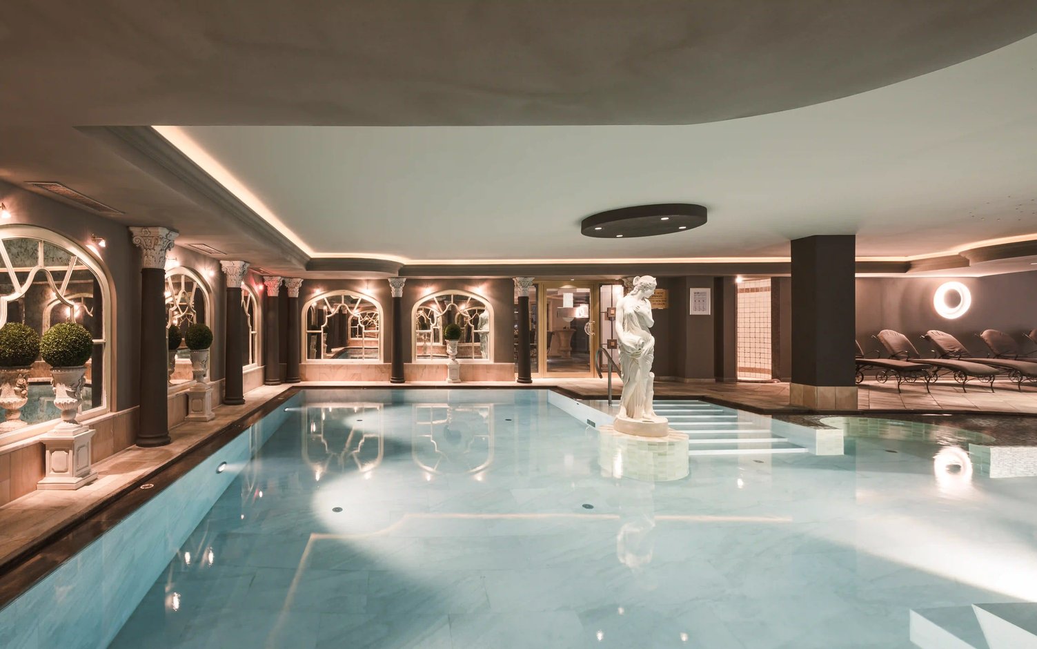 Eleganter Innenpool im Spa Bereich mit ruhigem Wasser, klassischer Statue im Becken, warmem Licht, Saeulen, Spiegelboegen und Liegen zur Entspannung